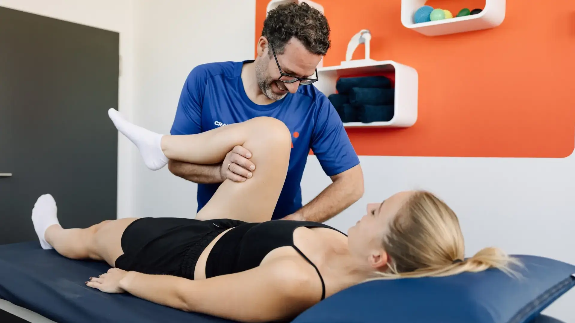 Physiotherapeut führt lächelnd eine Mobilisation des Beins bei einer liegenden Patientin in einem hellen Behandlungsraum durch.