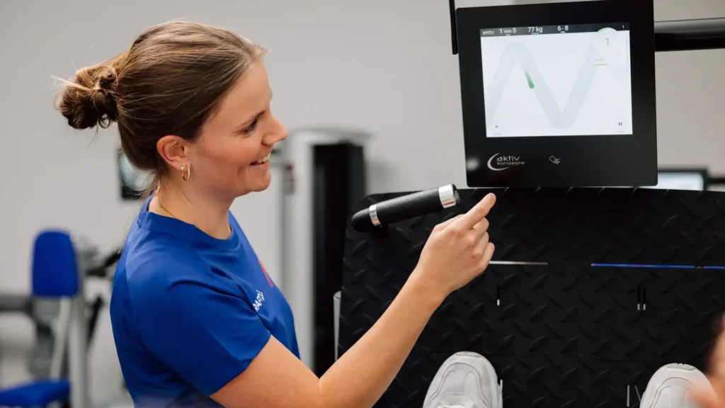 Physiotherapeutin erklärt einem Patienten die Anzeige auf dem Bildschirm eines Trainingsgeräts und begleitet seine Übung.