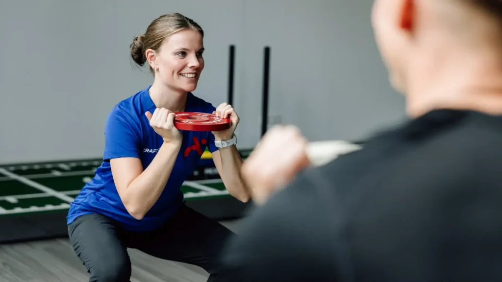 Physiotherapeutin zeigt Kniebeugenübung mit Gewichtsscheibe während eines Trainings in der Praxis.