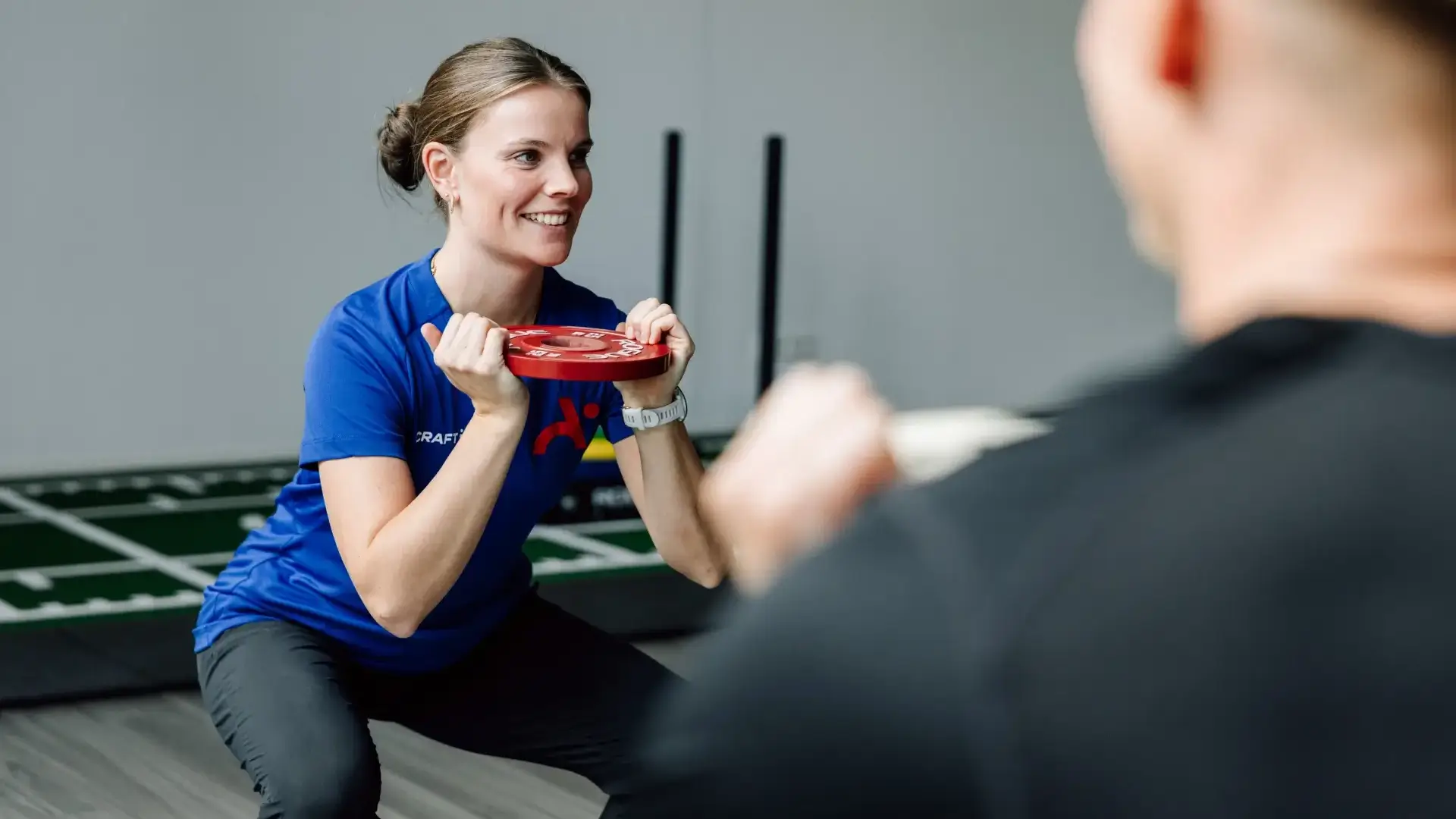 Physiotherapeutin zeigt Kniebeugenübung mit Gewichtsscheibe während eines Trainings in der Praxis.