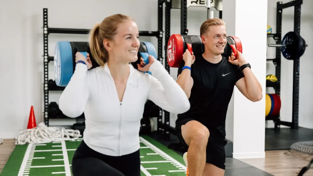Zwei Personen trainieren mit Sandsäcken und machen Ausfallschritte im Trainingsraum der Praxis.