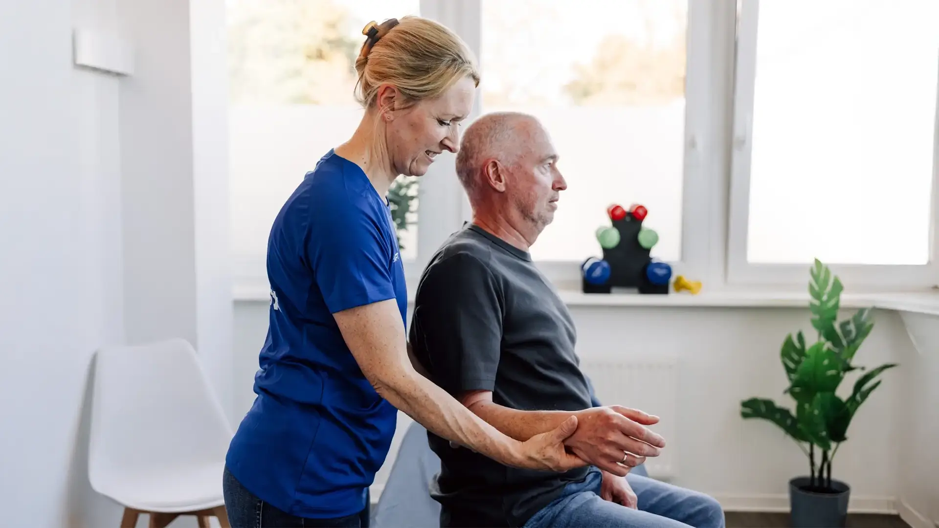 Physiotherapeutin unterstützt Patienten beim Aufrichten und bei einer Armübung im Sitzen.