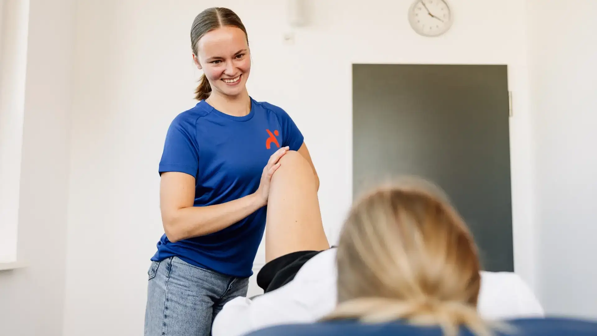Physiotherapeutin behandelt lächelnd das Knie einer Patientin auf der Liege.
