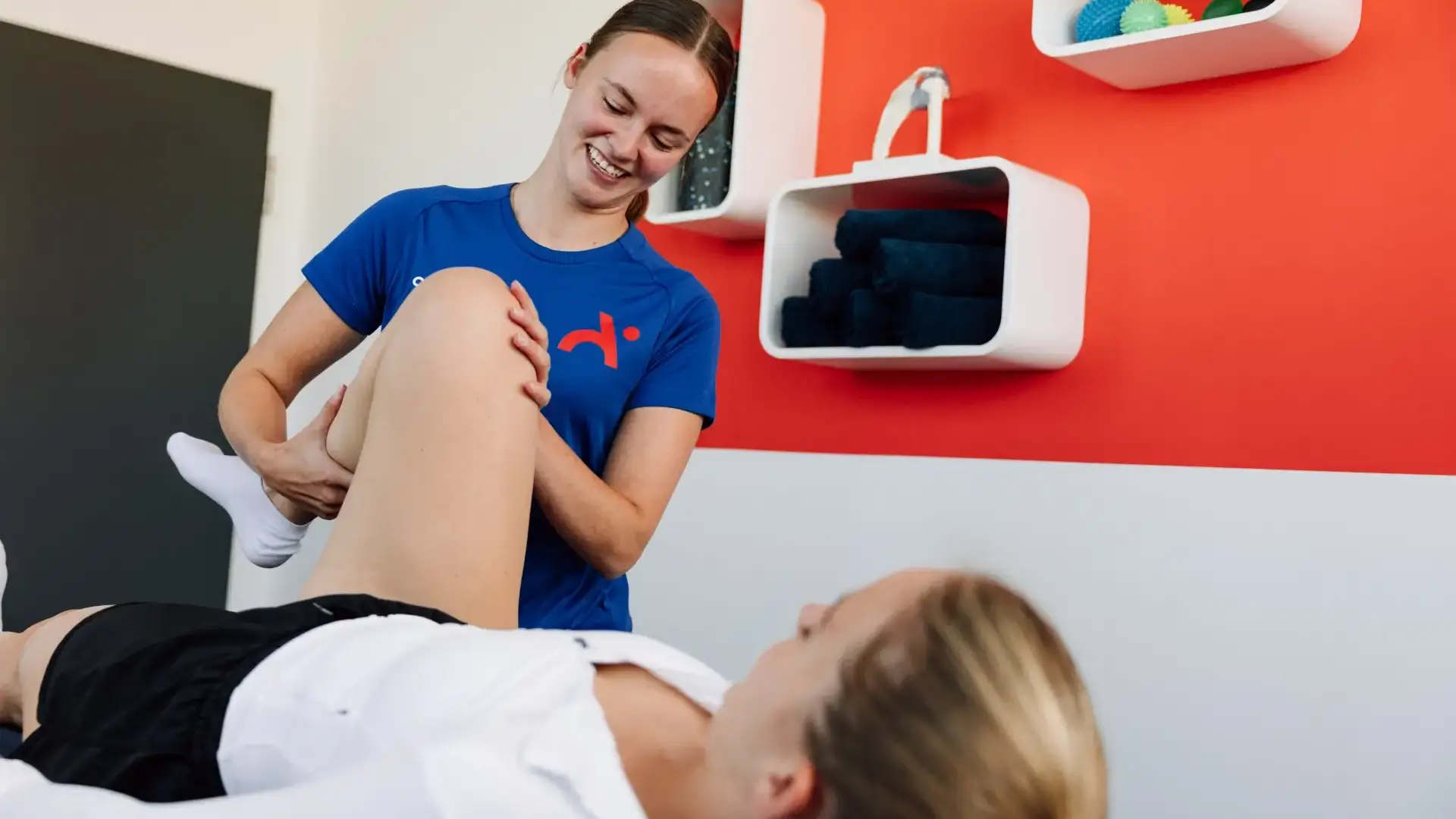 Physiotherapeutin mobilisiert das Bein einer liegenden Patientin vor oranger Praxiswand.