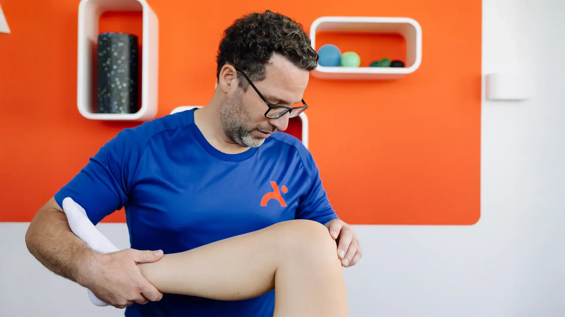 Physiotherapeut untersucht Kniegelenk einer Patientin vor oranger Praxiswand.