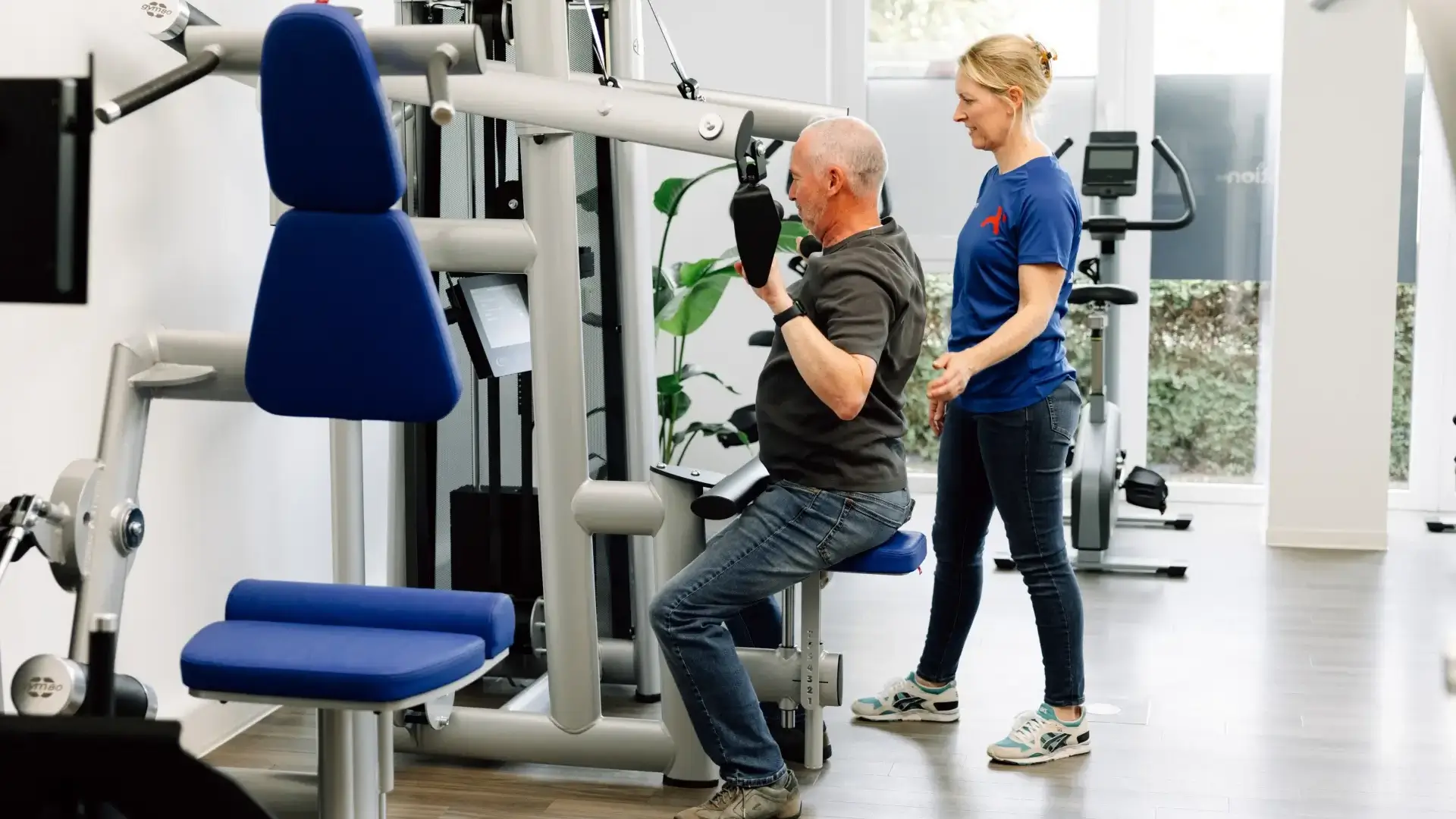 Physiotherapeutin betreut älteren Patienten beim Training an der Kraftmaschine in der Praxis.