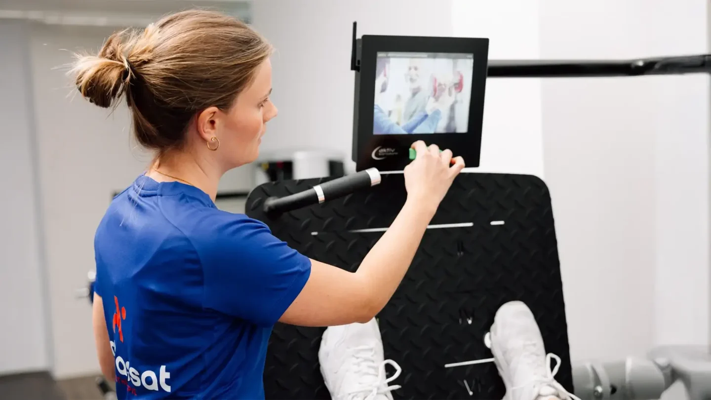 Physiotherapeutin stellt digitales Trainingsgerät ein, während Patientin an der Beinpresse trainiert.