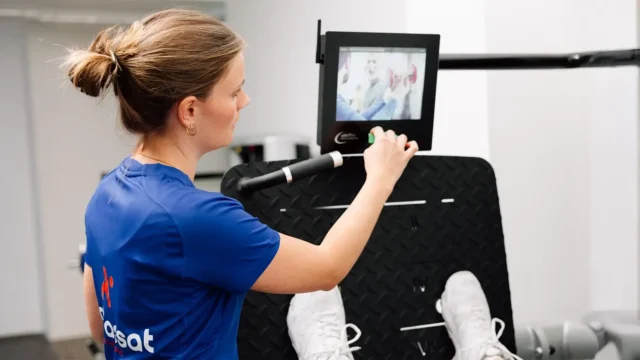 Physiotherapeutin stellt digitales Trainingsgerät ein, während Patientin an der Beinpresse trainiert.