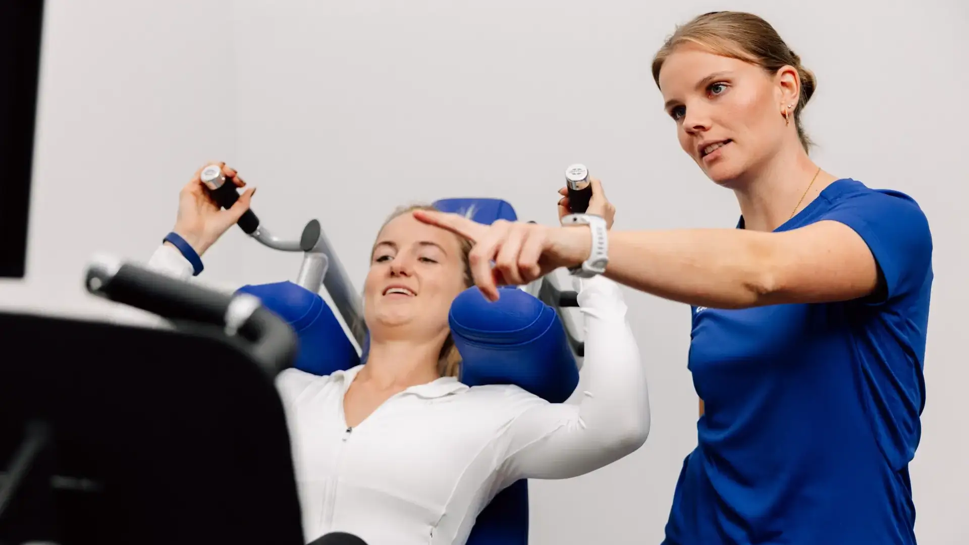 Physiotherapeutin weist Patientin bei einer Übung an der Schultermaschine im Trainingsraum an.