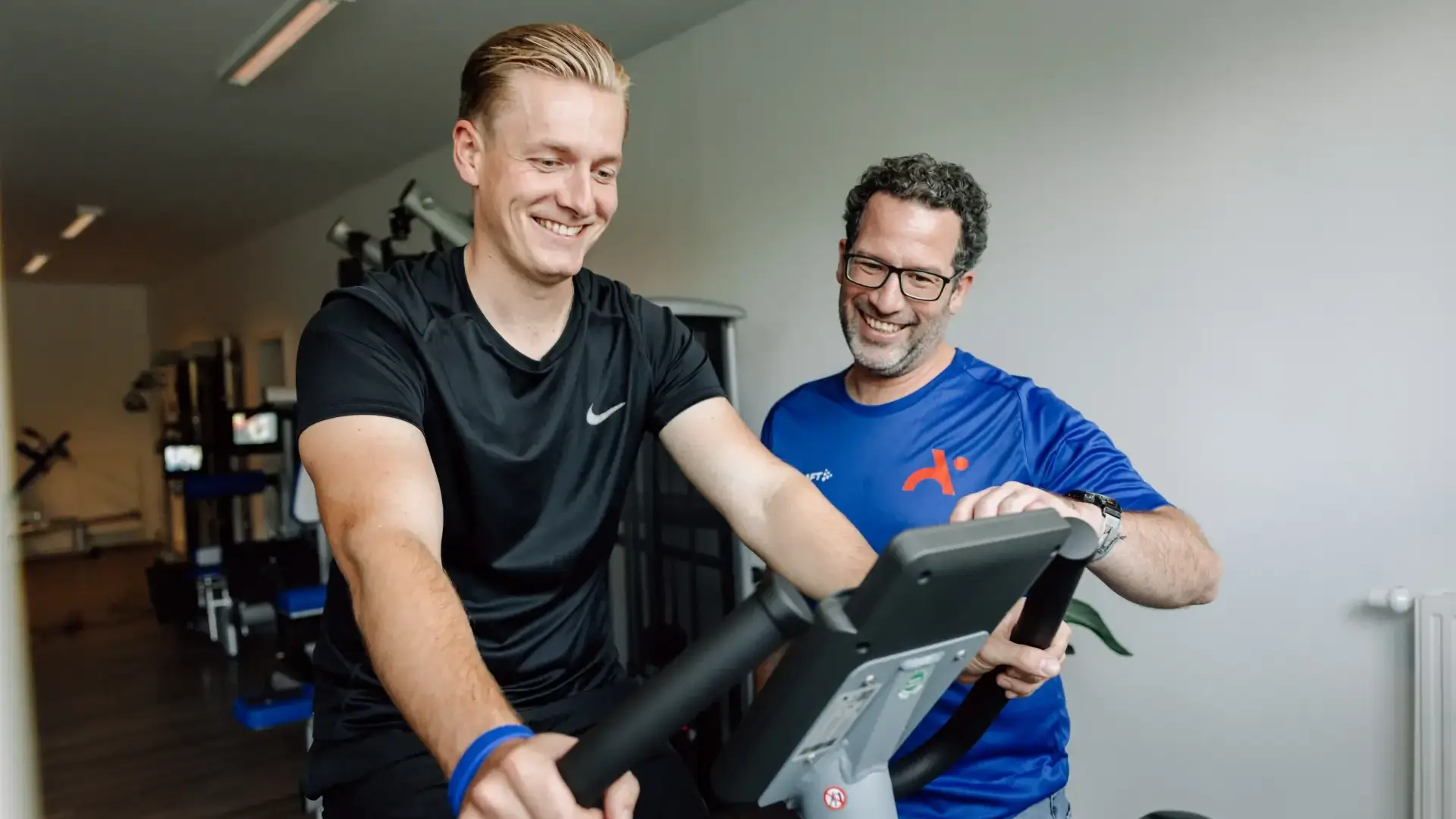 Patient fährt auf dem Fahrradergometer, betreut von Physiotherapeut in blauem Shirt.