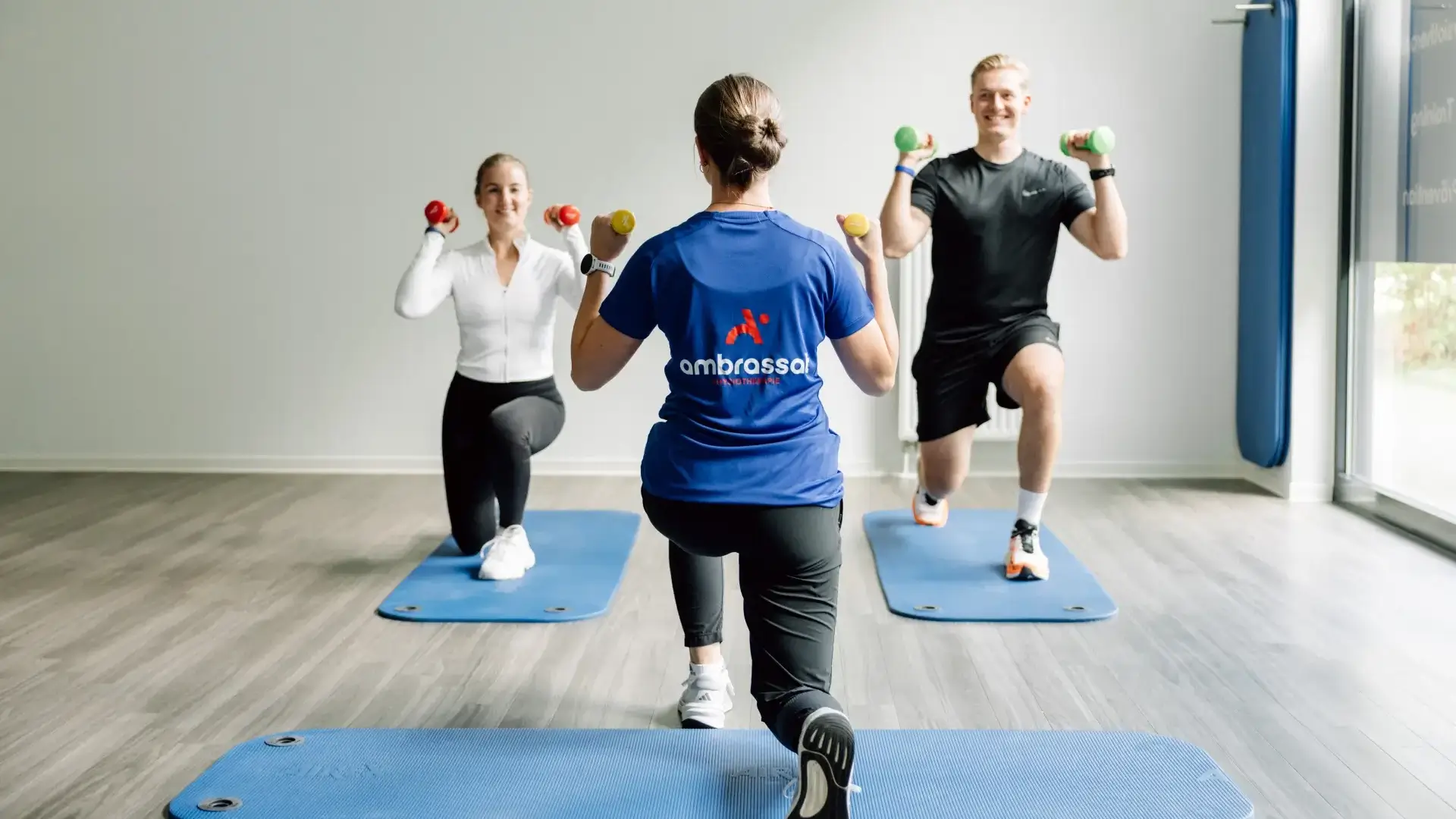 Physiotherapeutin leitet zwei Patientinnen bei Ausfallschritten mit Kurzhanteln auf Matten an.