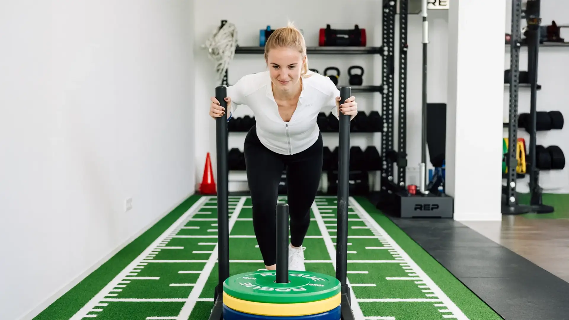 Frau führt Krafttraining mit Gewichtsschlitten auf Kunstrasen im Trainingsraum durch.
