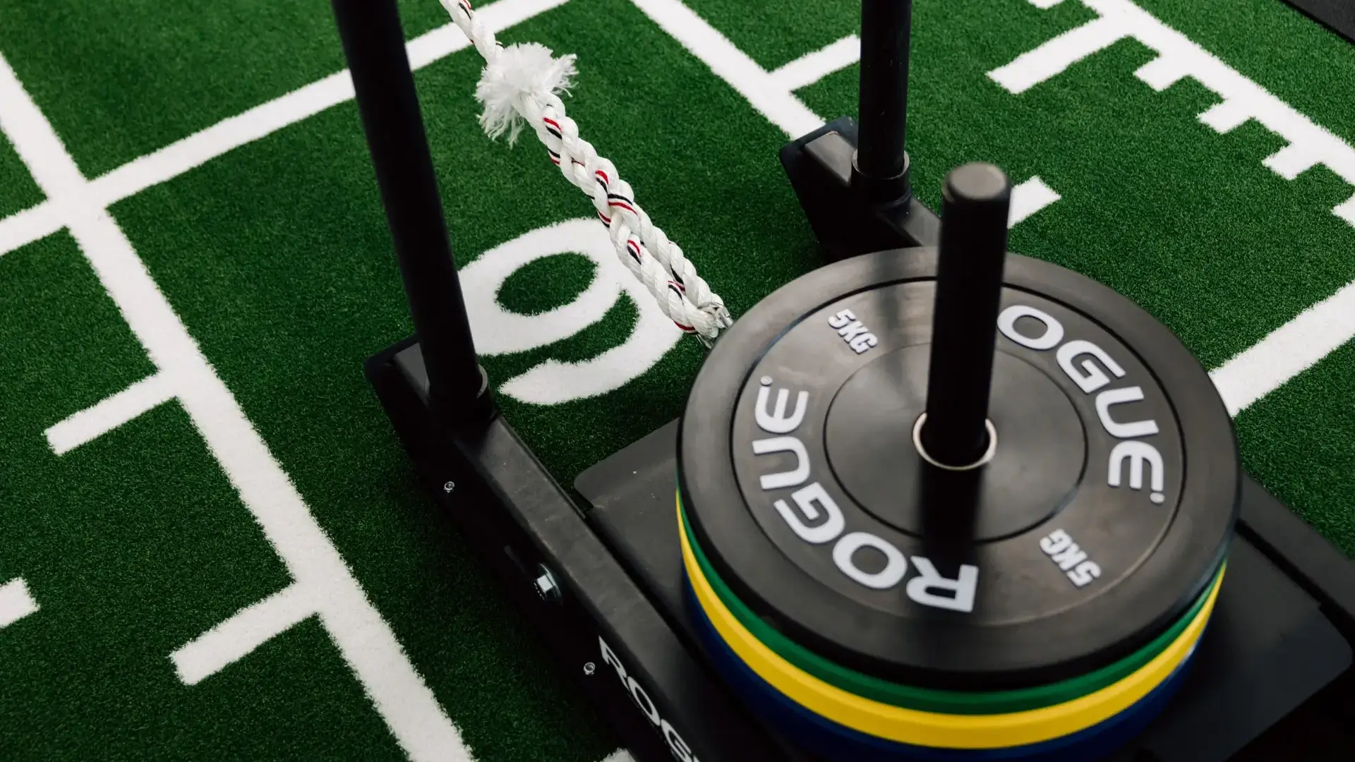 Detailaufnahme eines Gewichtsschlittens mit Hantelscheiben auf grünem Kunstrasen.