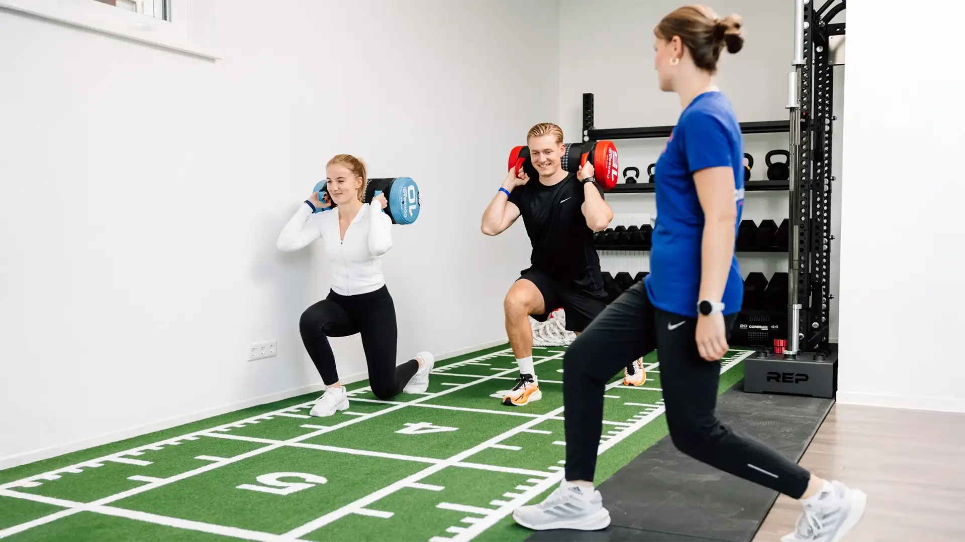 Zwei Personen führen Ausfallschritte mit Sandsäcken unter Anleitung einer Physiotherapeutin durch.