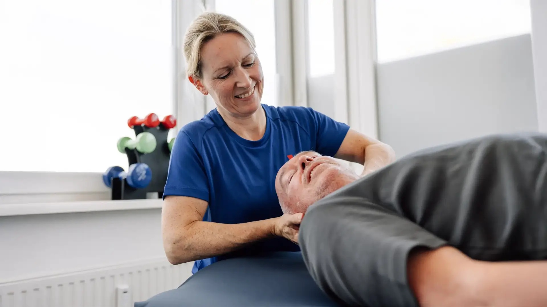 Physiotherapeutin behandelt den Kiefer- und Nackenbereich eines liegenden Patienten, sanfte manuelle Therapie im Behandlungsraum.